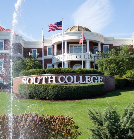 A brick building with the name "South College" displayed prominently on a curved sign in front. The structure has a dome and multiple flags, including the American flag, flying above. The lush lawn and a spraying fountain add to the picturesque campus setting.