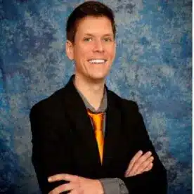 A person with short hair, glasses, and earrings is smiling with arms crossed. They are wearing a black sweater over a maroon top in front of a blue textured background.