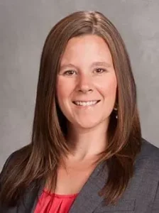 Breanna (Bre) Reynolds A woman with long, straight brown hair smiles at the camera. She is wearing a gray blazer over a pink top, and the background is a plain gray backdrop.
