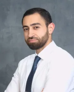 Ahmad Sabbahi, PT, MA, CCS A man with short dark hair and a neatly trimmed beard is standing against a gray backdrop. He is wearing a white collared shirt and a patterned blue tie, looking at the camera with a neutral expression.