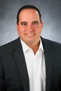 A man with short dark hair, wearing a white shirt and a dark grey blazer, posed against a grey studio backdrop. He is smiling at the camera.