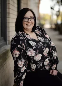 c.siracusa A woman with shoulder-length dark hair and glasses is smiling while leaning against a brick wall outdoors. She is wearing a black floral blouse.