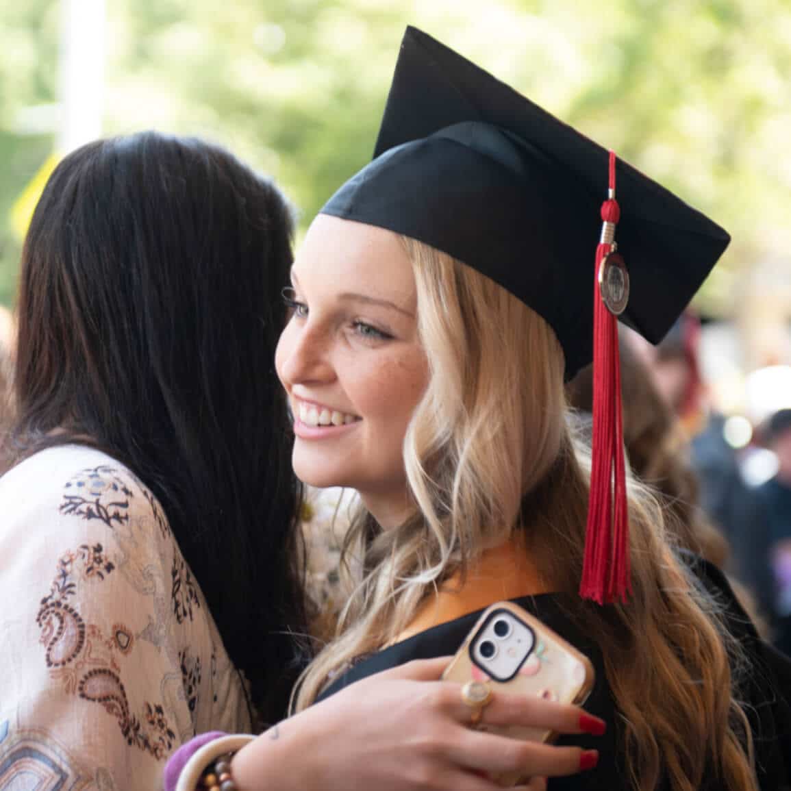 A woman wearing a graduation cap with a red tassel smiles as she hugs someone whose back is to the camera.