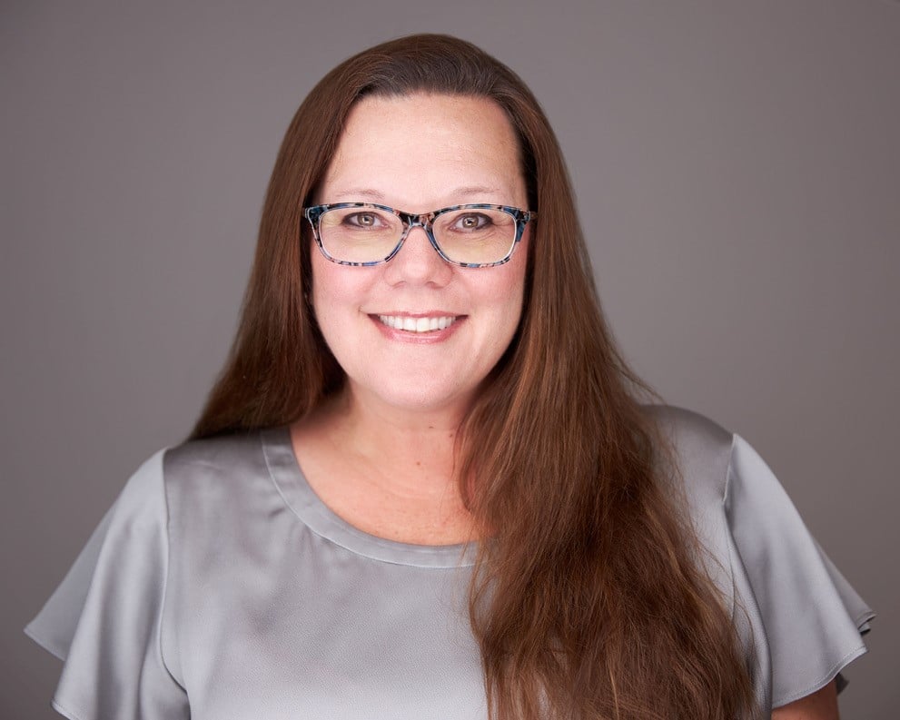 A woman with long brown hair and glasses is smiling at the camera. She is wearing a grey top and stands against a plain grey background.