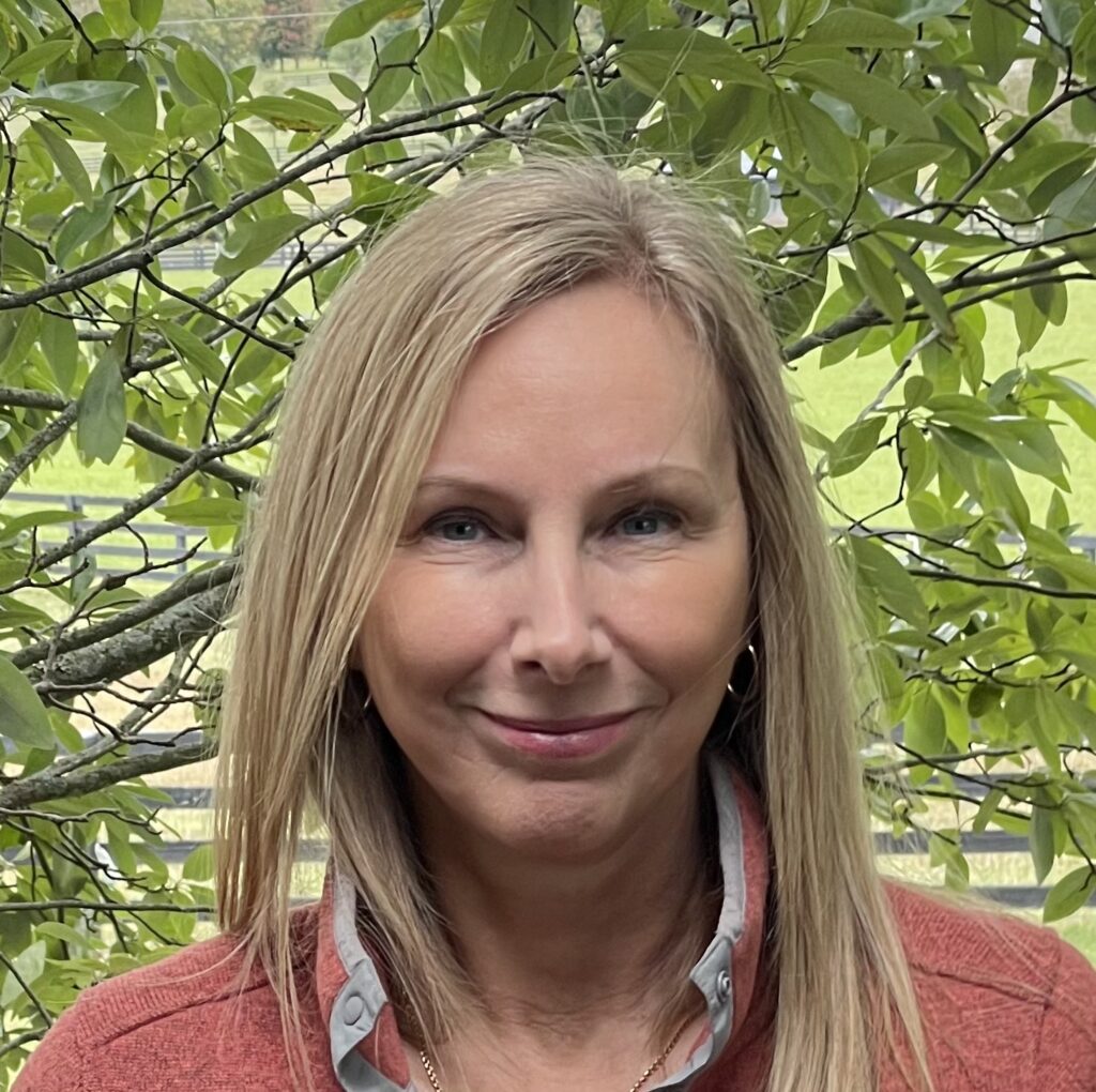 A woman with light blonde hair and a light complexion is smiling. She is wearing a coral-colored top and standing in front of green foliage with branches, giving the photo a natural outdoor setting.