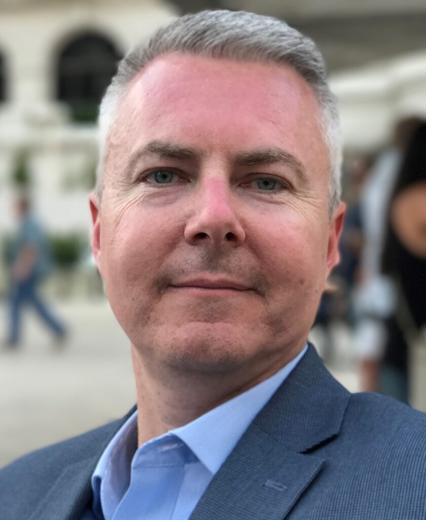 A close-up of a man with short gray hair and a trimmed beard, wearing a blue dress shirt and gray blazer. He is looking directly at the camera with a slight smile. The background is blurred with people and a building with arches in soft focus.