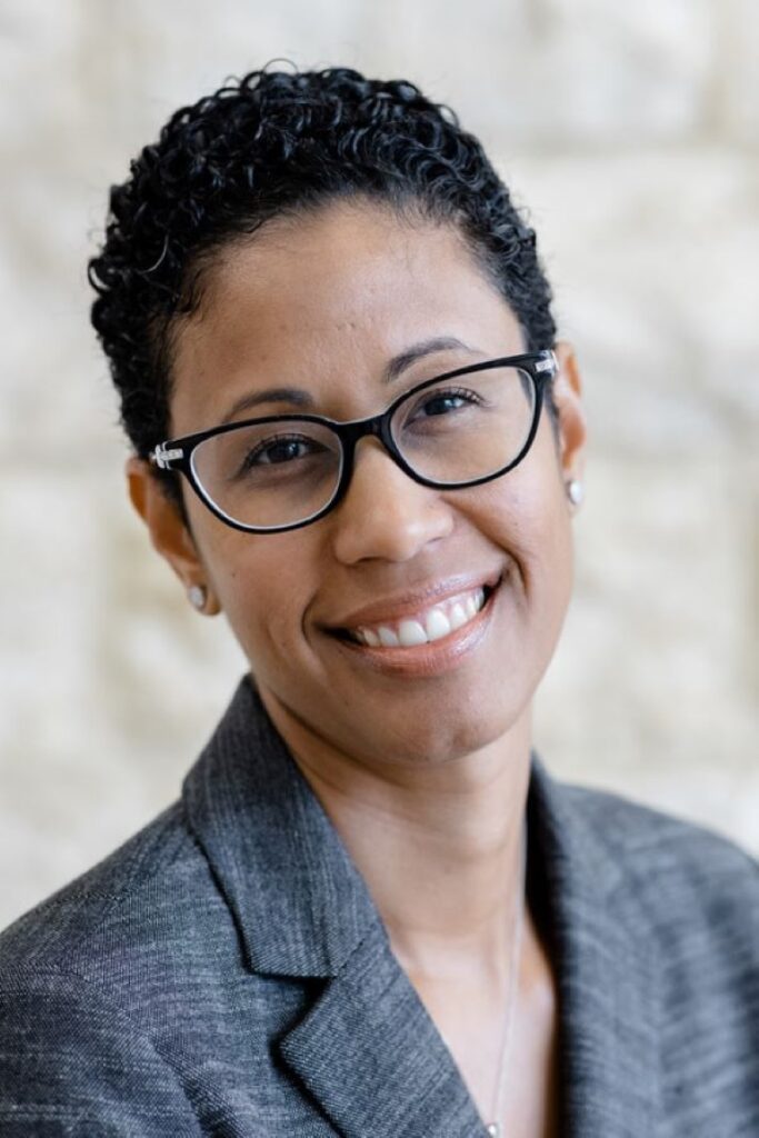 A person with short curly hair and black-rimmed glasses is smiling at the camera. They are wearing a grey blazer and a white shirt, with a light-colored background behind them.