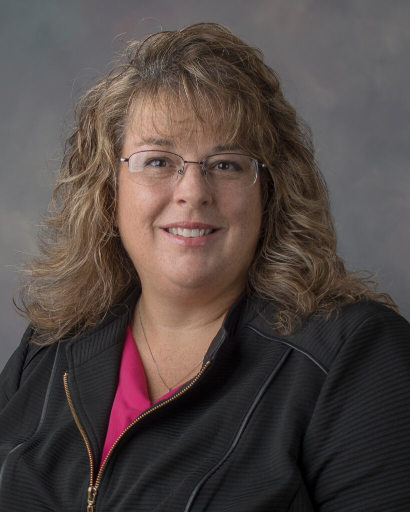 A woman with curly, shoulder-length blonde hair and glasses smiles at the camera. She is wearing a black jacket over a pink top, and the background is a neutral gray.