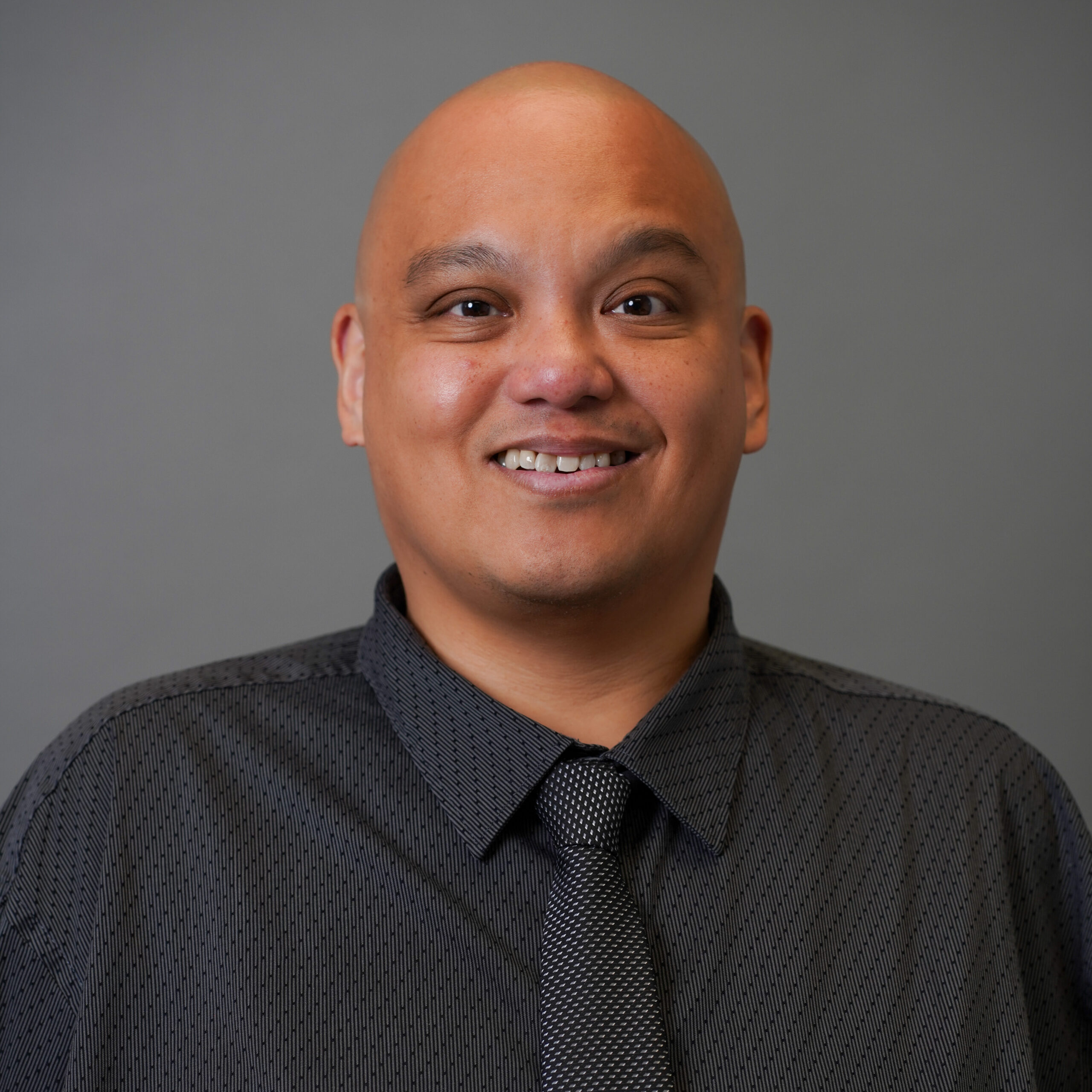 A man with a shaved head smiles warmly at the camera. He is wearing a dark gray, patterned button-up shirt with a matching tie. The background is solid gray.