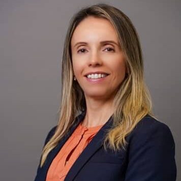 A woman with long, straight blonde hair is smiling at the camera. She is wearing a navy blazer over an orange blouse and is standing against a plain grey background.
