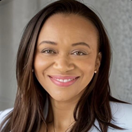 A woman with long, straight hair is smiling warmly. She is wearing a light-colored blazer and earrings. The background appears to be an indoor setting with a gray wall.