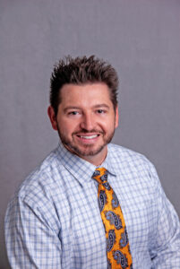 Miranda, Nicole A man with short, spiky hair and a trimmed beard smiles at the camera. He is wearing a light blue checkered dress shirt and an orange tie with blue paisley patterns. The background is plain and gray.