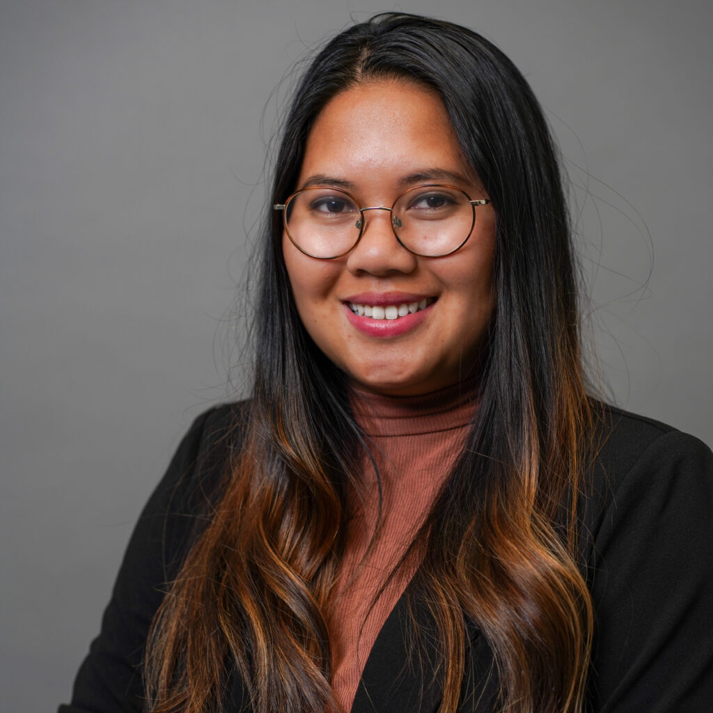 A woman with long, dark hair and glasses smiles at the camera. She is wearing a black blazer over a brown top. The background is a plain gray.