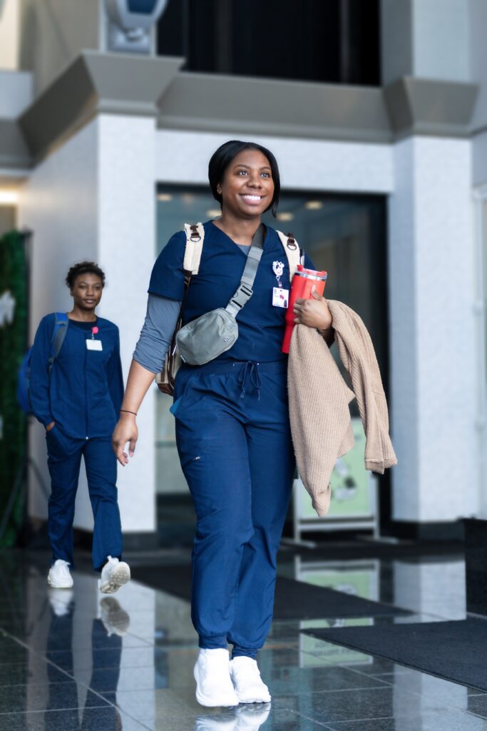 A student in navy blue scrubs walks through a building, holding a tan jacket and a red cup, with a gray crossbody bag. Another person in similar attire follows behind. Both wear ID badges and appear to be smiling, possibly discussing their experiences applying for the Tennessee Promise Scholarship.