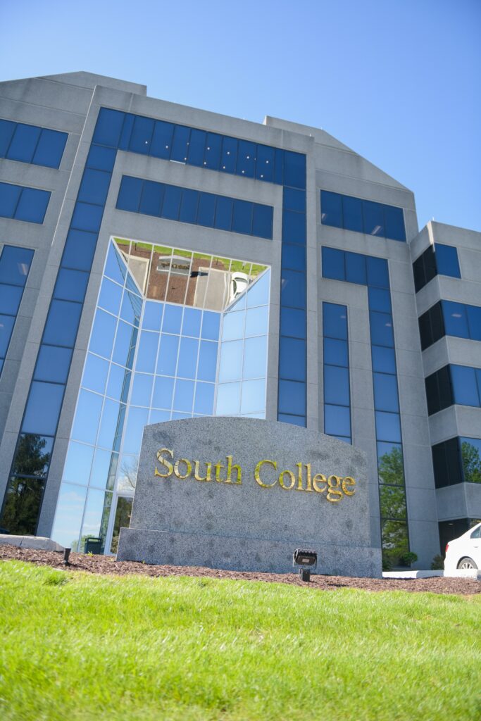 A modern building with numerous windows, labeled "South College." In front of the building is a large, gray stone sign with gold lettering that also reads "South College." The building is surrounded by a well-manicured green lawn under a clear blue sky.