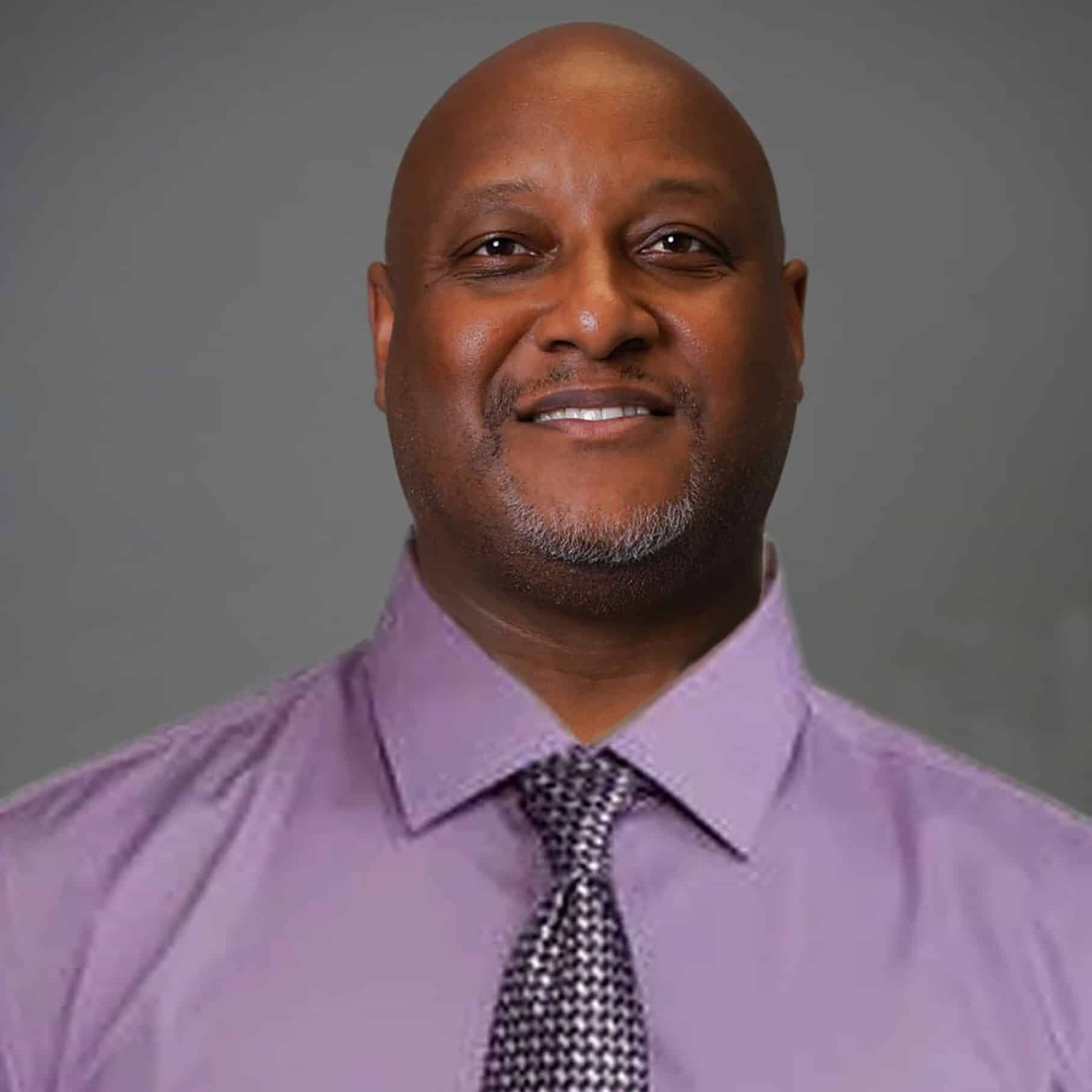 A man with a shaved head and goatee is smiling at the camera. He is wearing a light purple collared shirt and a patterned tie, standing against a plain gray background.