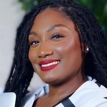 A smiling person with long, dark, braided hair and red lipstick is pictured. They are wearing a white top with black trim. The background is blurred greenery, suggesting an indoor or outdoor setting.