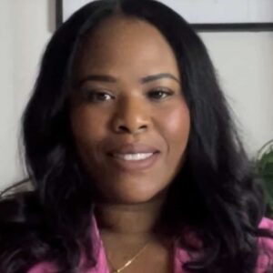 A woman with long, black hair is smiling at the camera. She is wearing a pink top and a necklace. The background is partially visible with a plant and a framed picture.