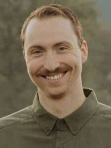 A person with short, red hair and a mustache is smiling. They are wearing a green button-up shirt with a collar. The background appears to be outdoors with greenery.