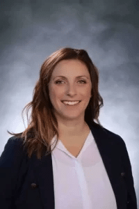 A woman with long, wavy brown hair is smiling at the camera. She is wearing a white blouse and a dark jacket. The background is a blurred gradient of gray tones.