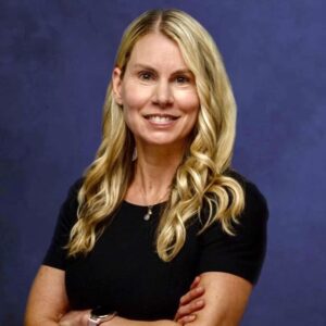 A blonde woman with long, wavy hair is standing against a purple backdrop, smiling. She is wearing a black top and has her arms crossed. A watch adorns her left wrist, and she has a necklace around her neck.