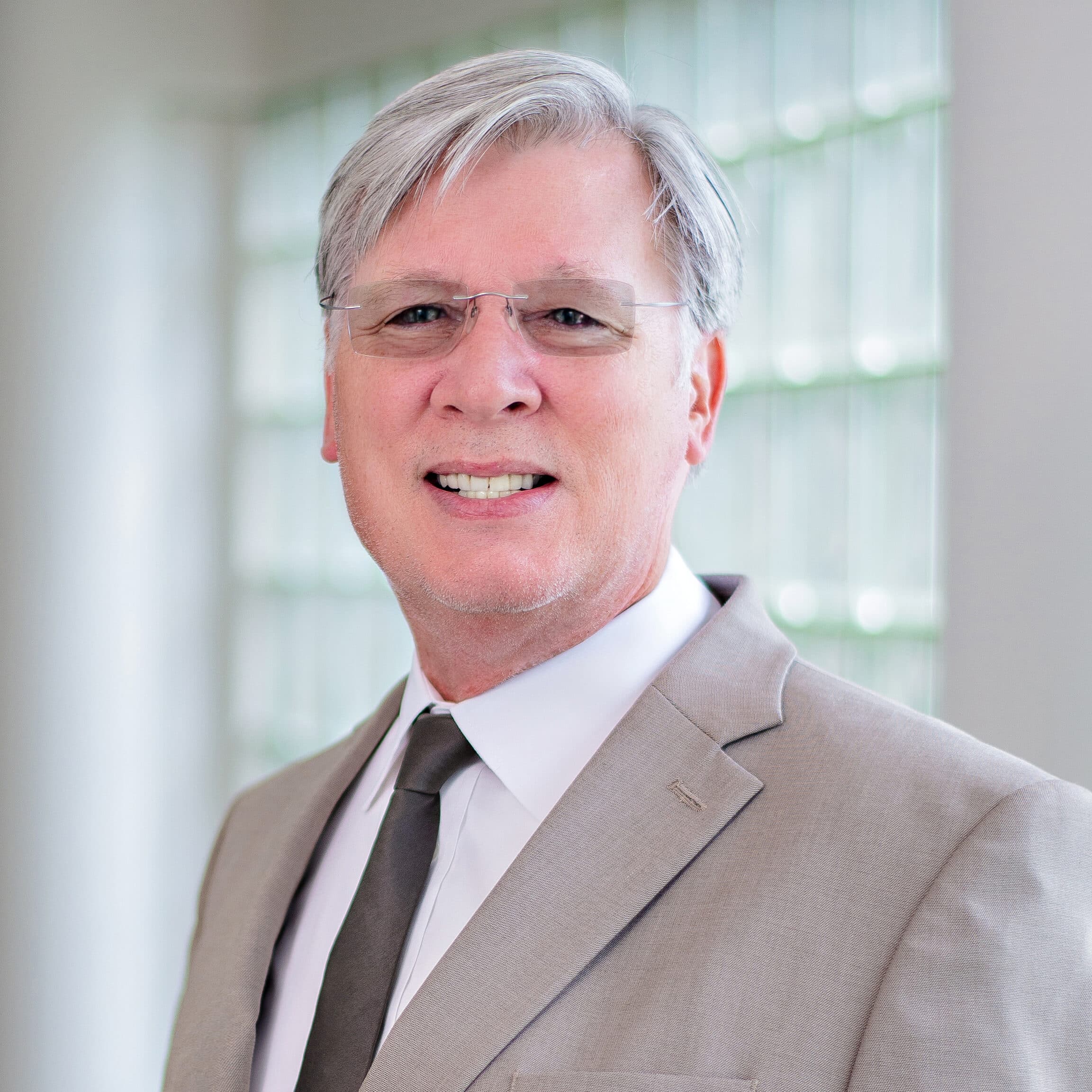 A person with short gray hair, glasses, and a light gray suit with a black tie is smiling at the camera. The background features a wall with rectangular glass blocks and is brightly lit.