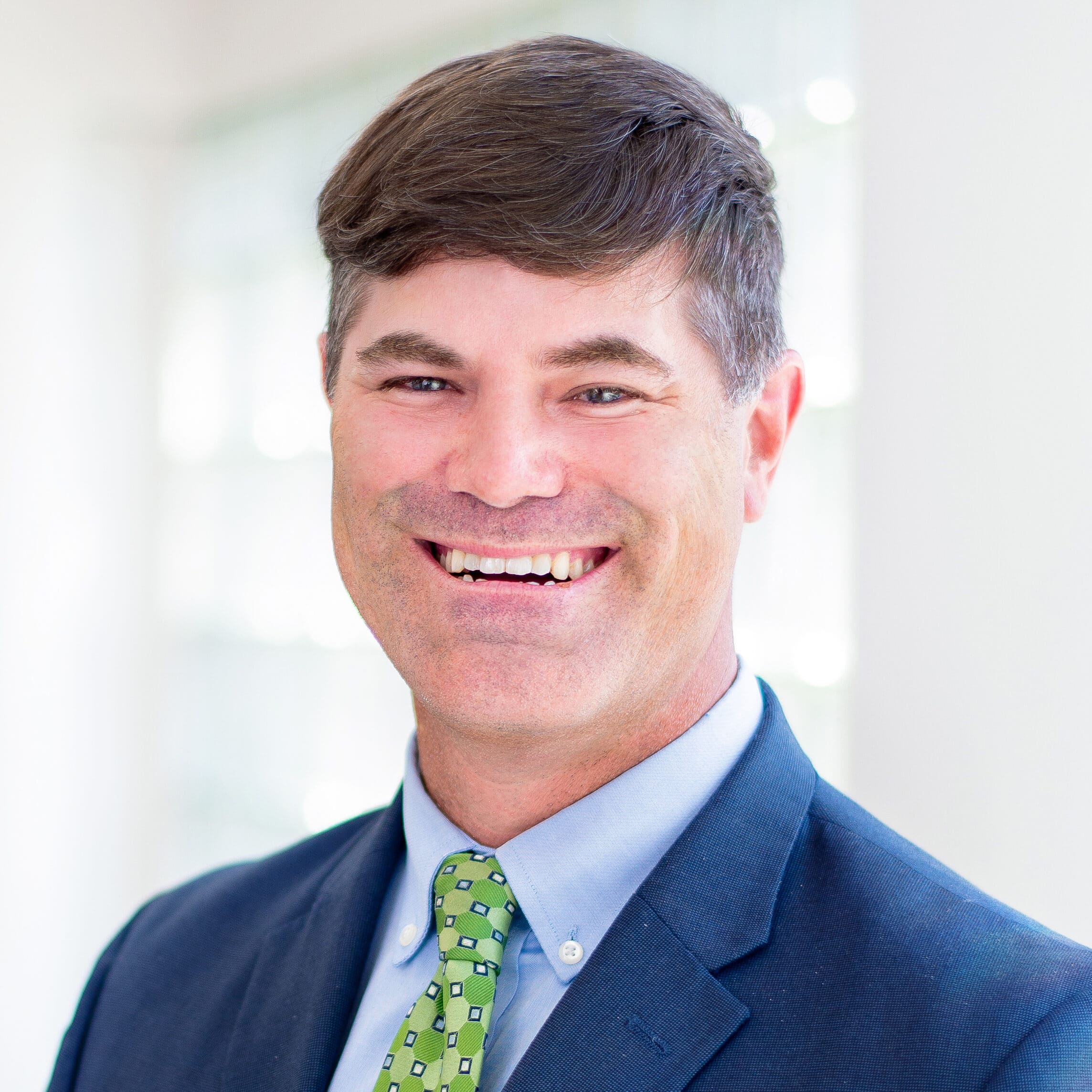 A smiling man with short brown hair is wearing a dark blue suit, light blue shirt, and a green tie with small white squares. He is standing in a brightly lit room with white walls and large windows in the background.
