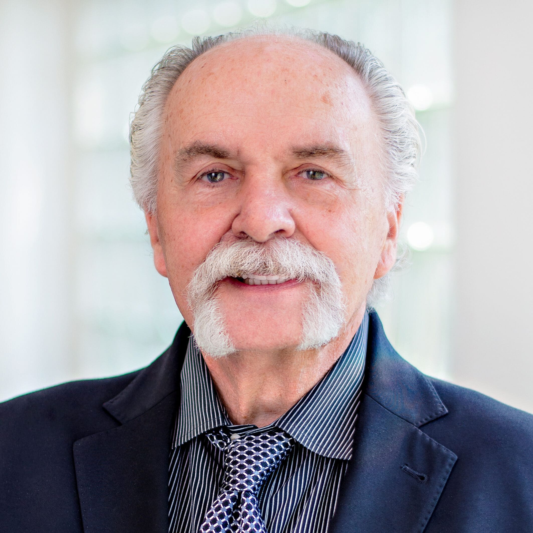 A man with gray hair and a mustache is smiling. He is wearing a dark suit jacket, a striped shirt, and a patterned tie. The background is softly blurred, suggesting an indoor setting with natural light.