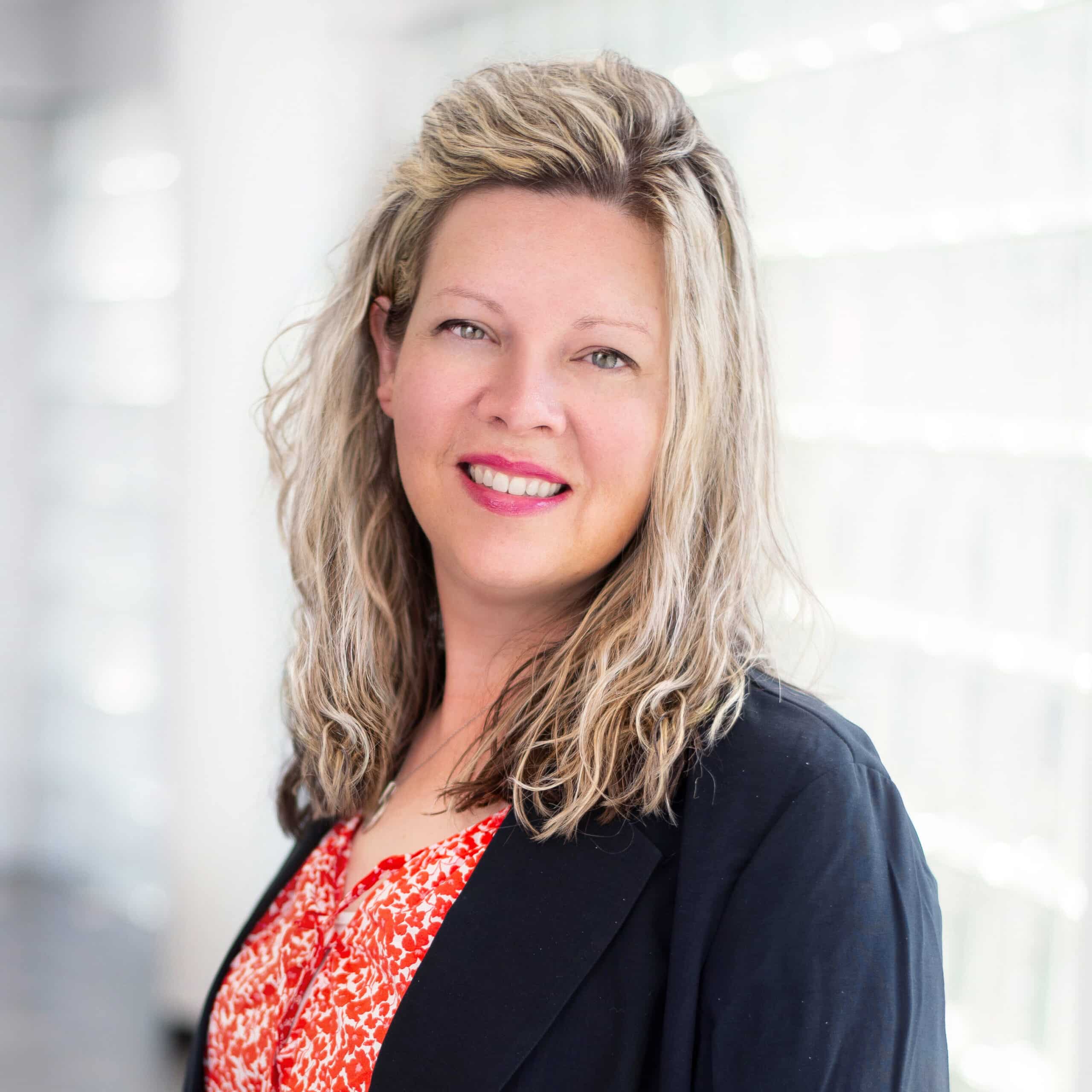 A woman with shoulder-length blonde hair stands indoors, smiling at the camera. She wears a red patterned blouse under a black blazer. The background is softly lit with large windows, giving a bright and airy feel.