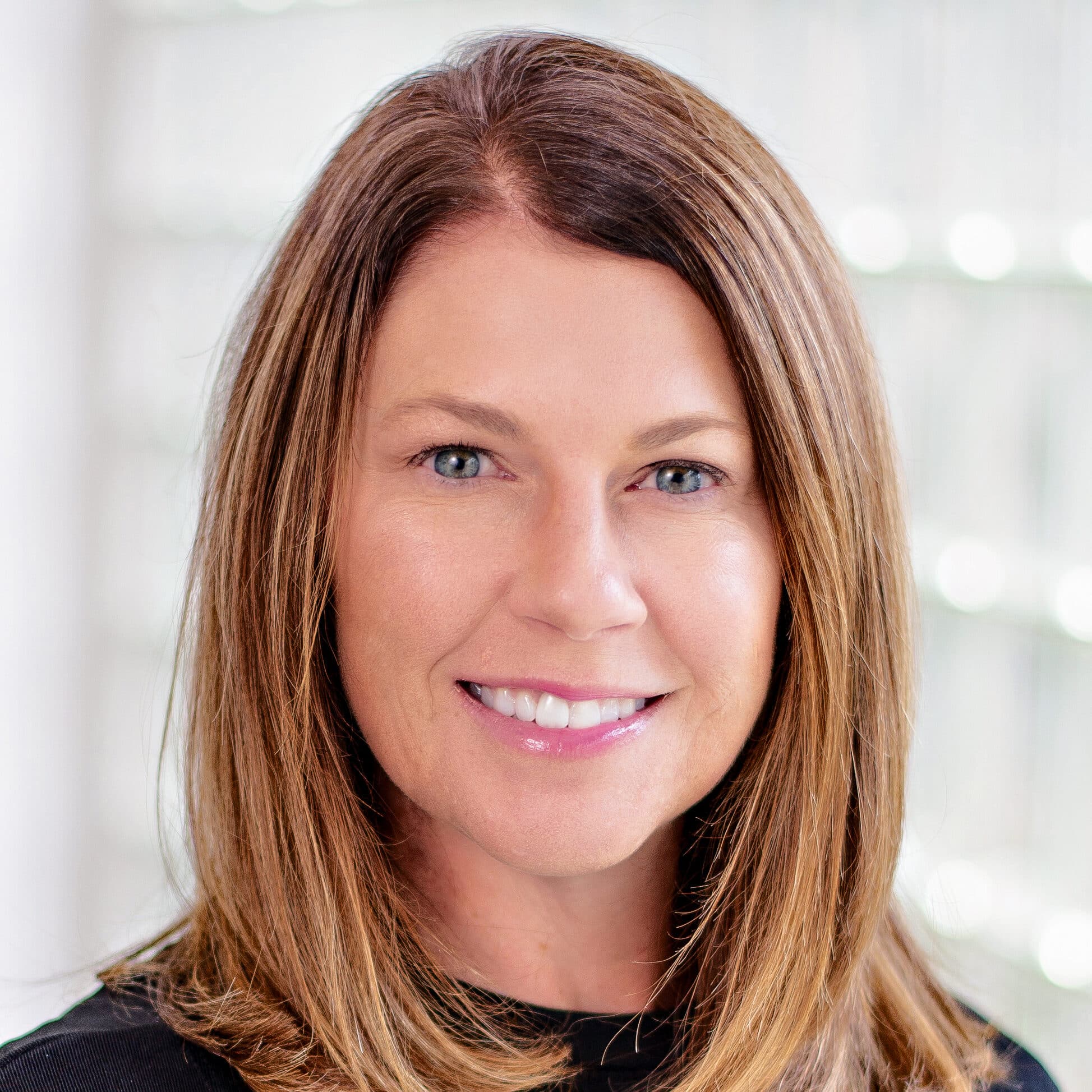 A woman with straight shoulder-length light brown hair and blue eyes smiling while wearing a black top. She stands against a softly blurred background with a light, airy feel.
