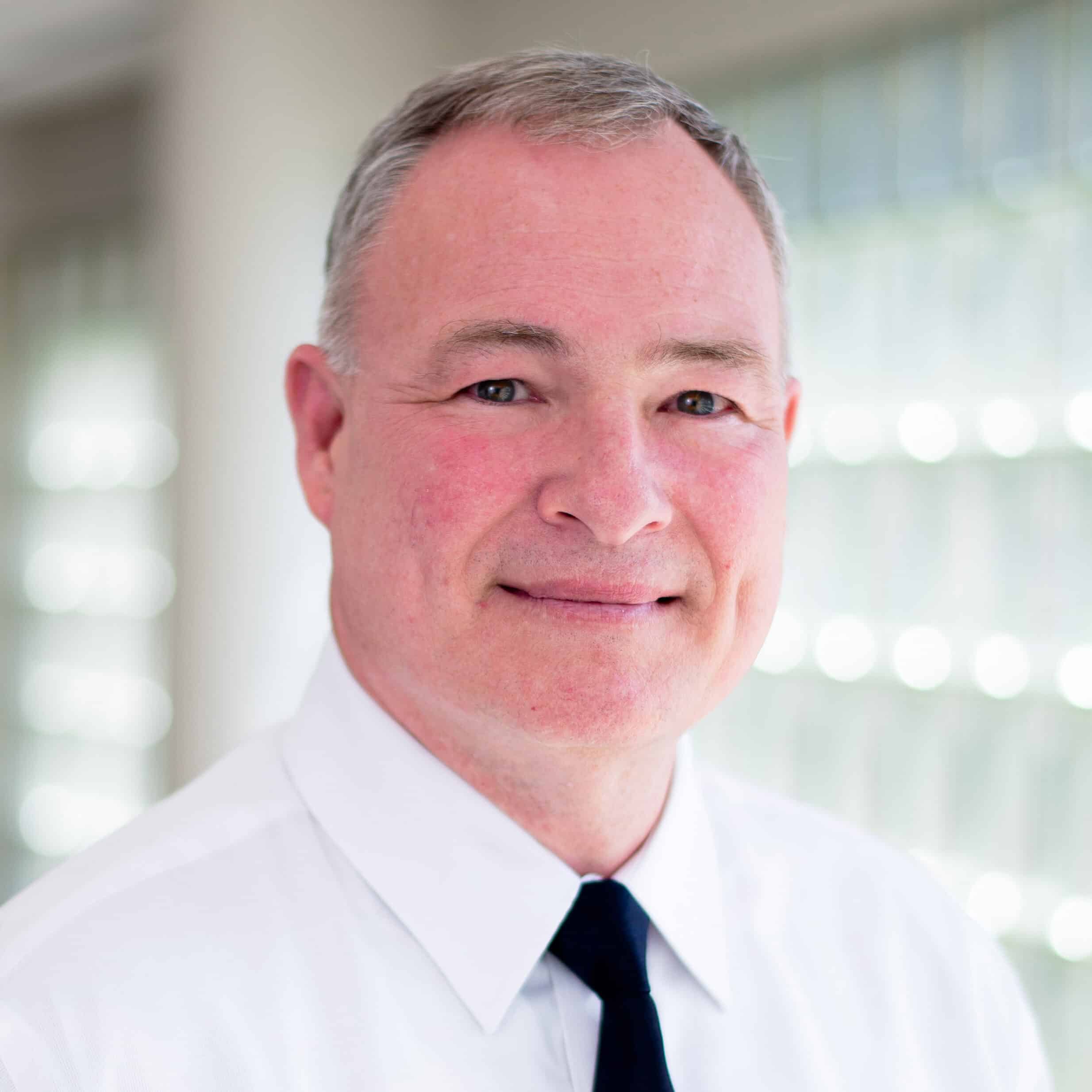 A man with short gray hair, smiling gently. He is wearing a white dress shirt and a dark tie, standing in front of a softly lit background.