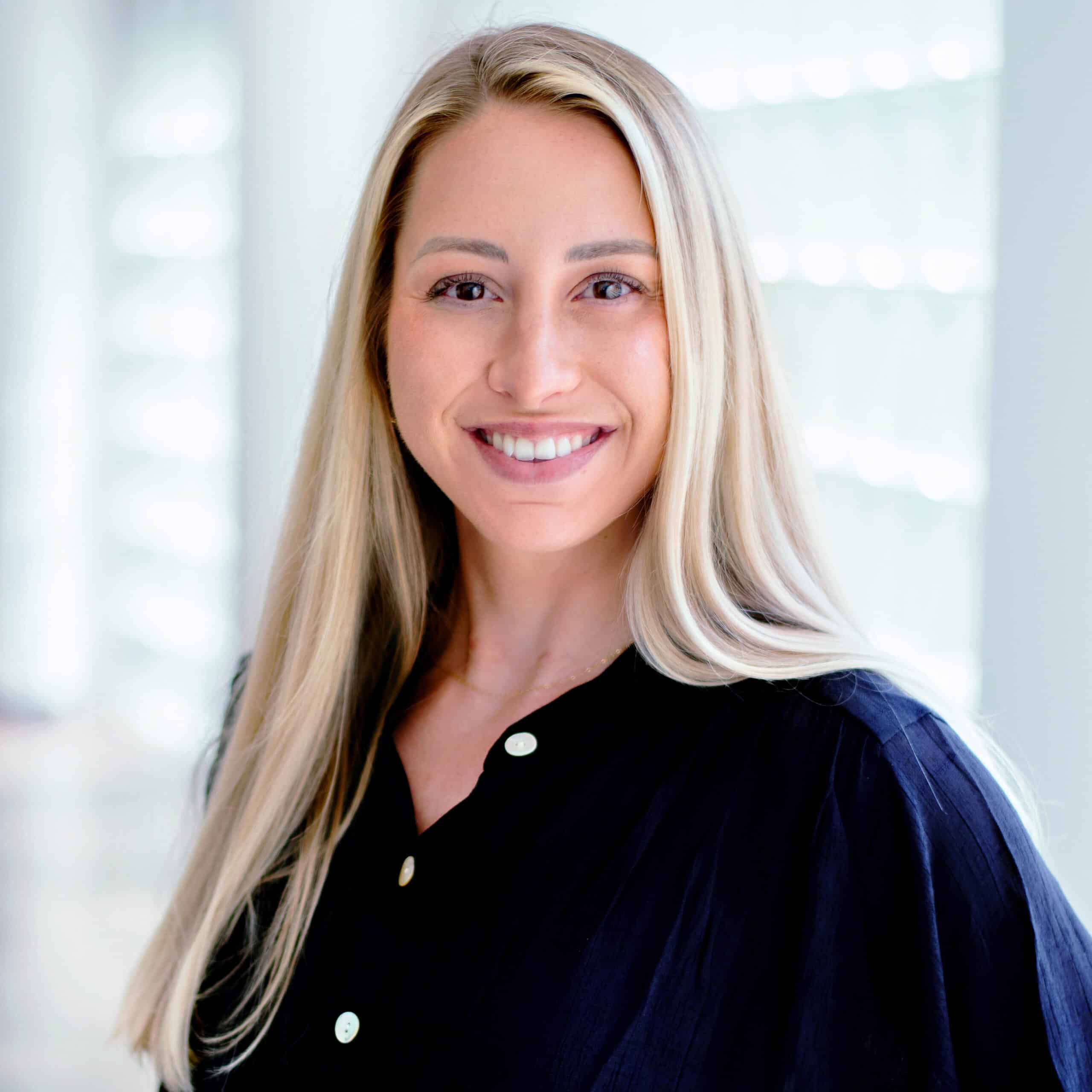 A person with long, straight blonde hair smiles warmly, wearing a dark button-up shirt. The background is softly blurred with light from windows, giving a bright and airy atmosphere.