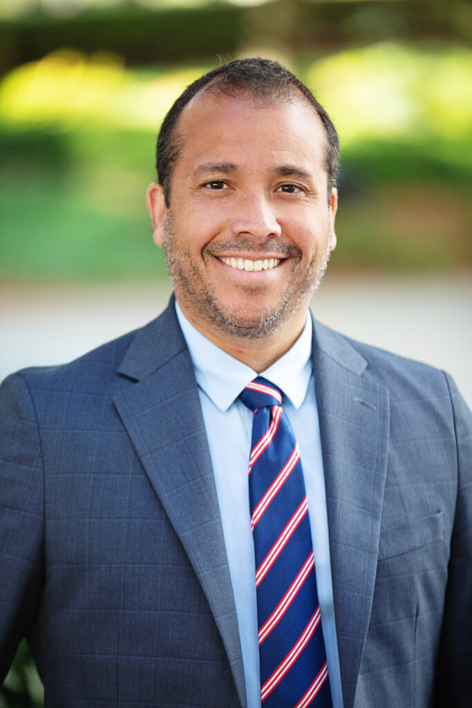A man with short dark hair and a beard smiles warmly at the camera. He is wearing a blue suit, a light blue shirt, and a blue tie with red and white stripes. The background is blurred with green foliage, suggesting an outdoor setting.
