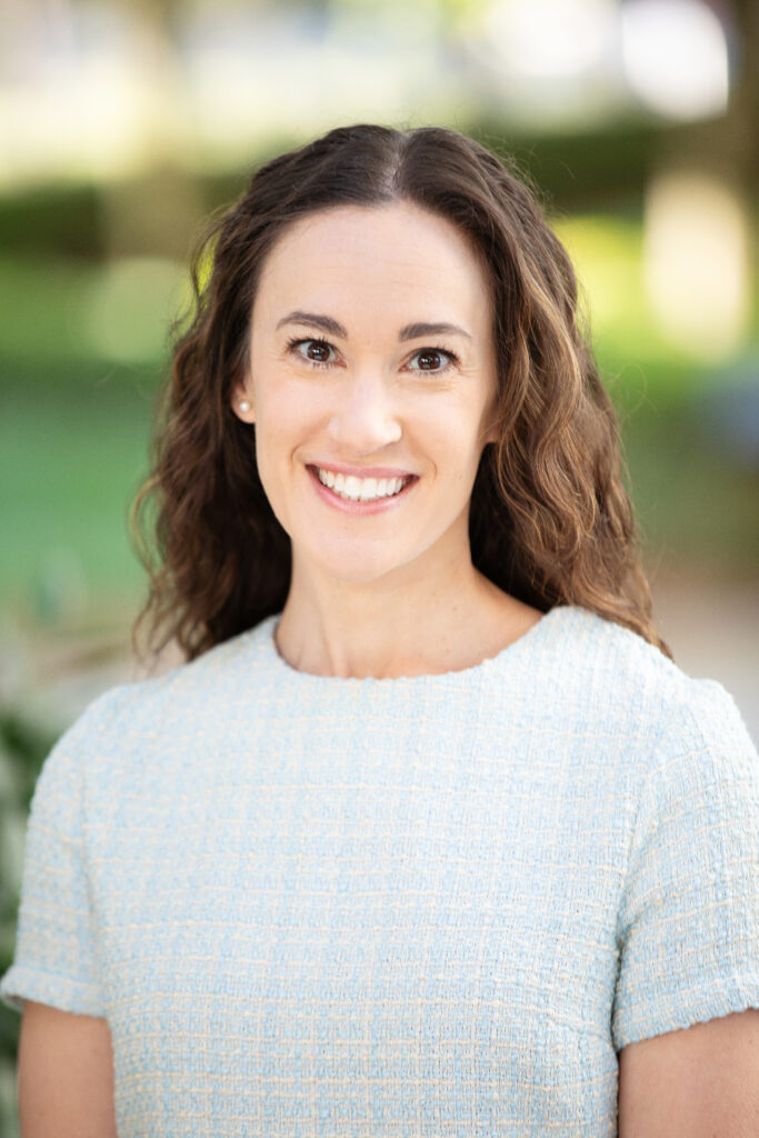 A woman with long, curly brown hair is smiling at the camera. She is wearing a light blue short-sleeved top and standing outdoors with a blurred green background.