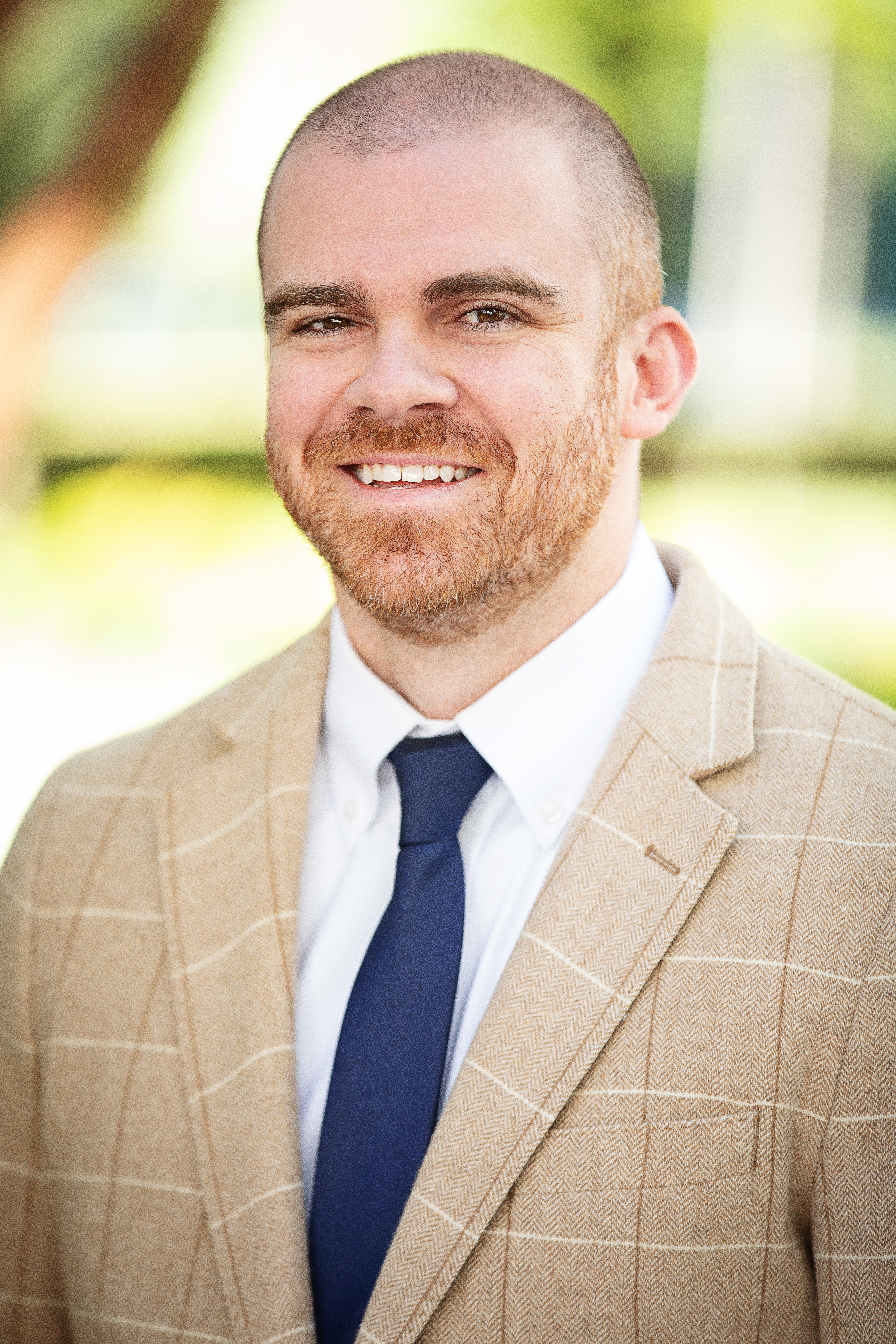 A man with a beard and a shaved head is wearing a beige plaid suit jacket, a white dress shirt, and a dark blue tie. He is smiling and the background is softly blurred with greenery.