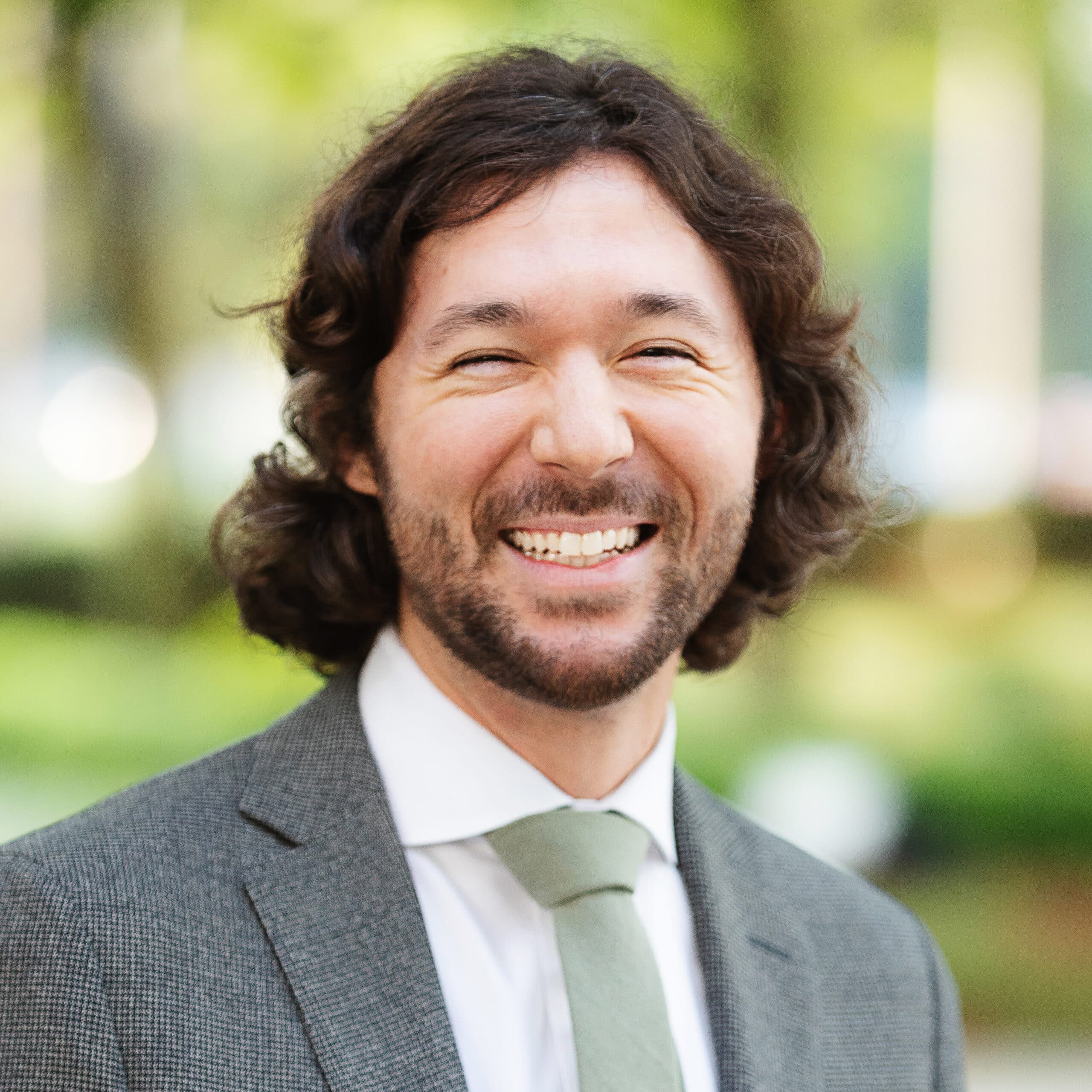 A man with medium-length curly hair and a beard is smiling broadly. He is wearing a grey suit jacket, white dress shirt, and a pale green tie.