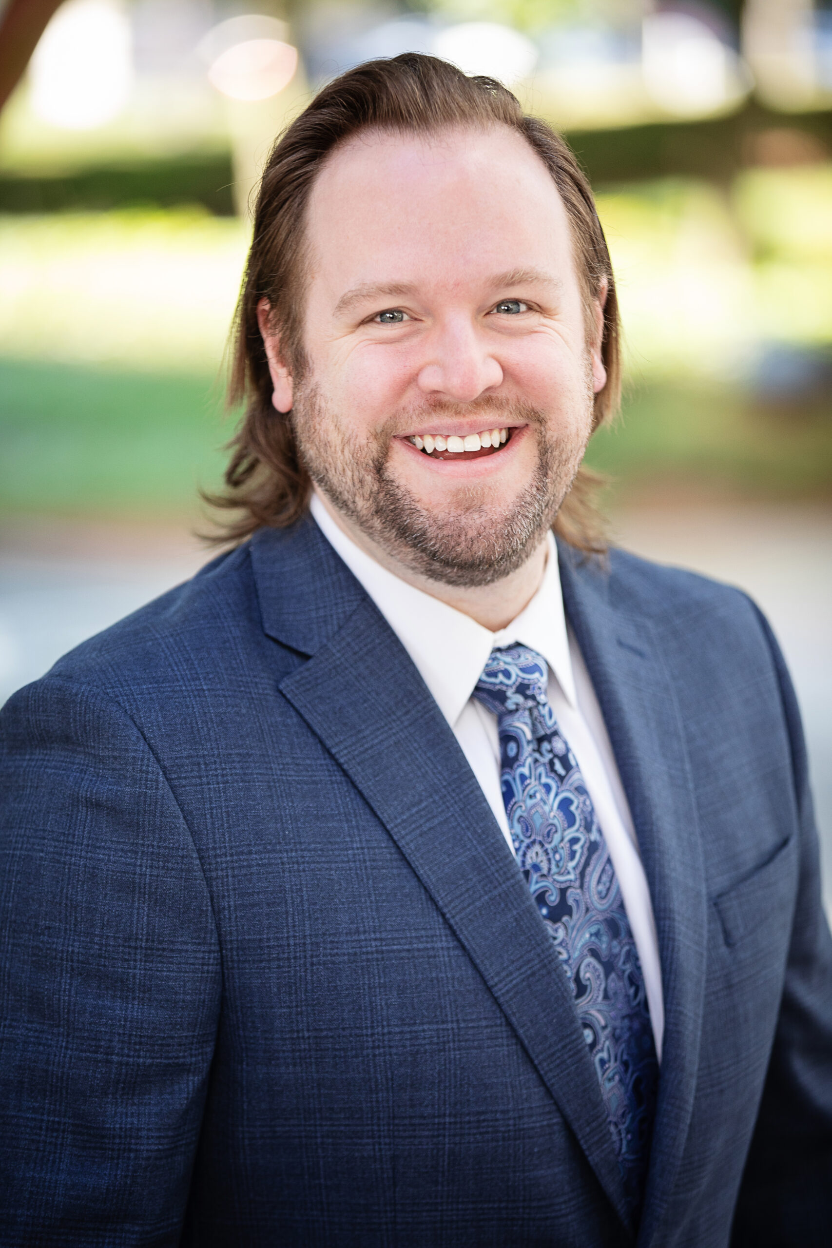 A bearded man with shoulder-length hair smiles warmly at the camera. He is wearing a blue suit jacket, white dress shirt, and a blue paisley tie. The background is blurred with greenery and outdoor elements visible.