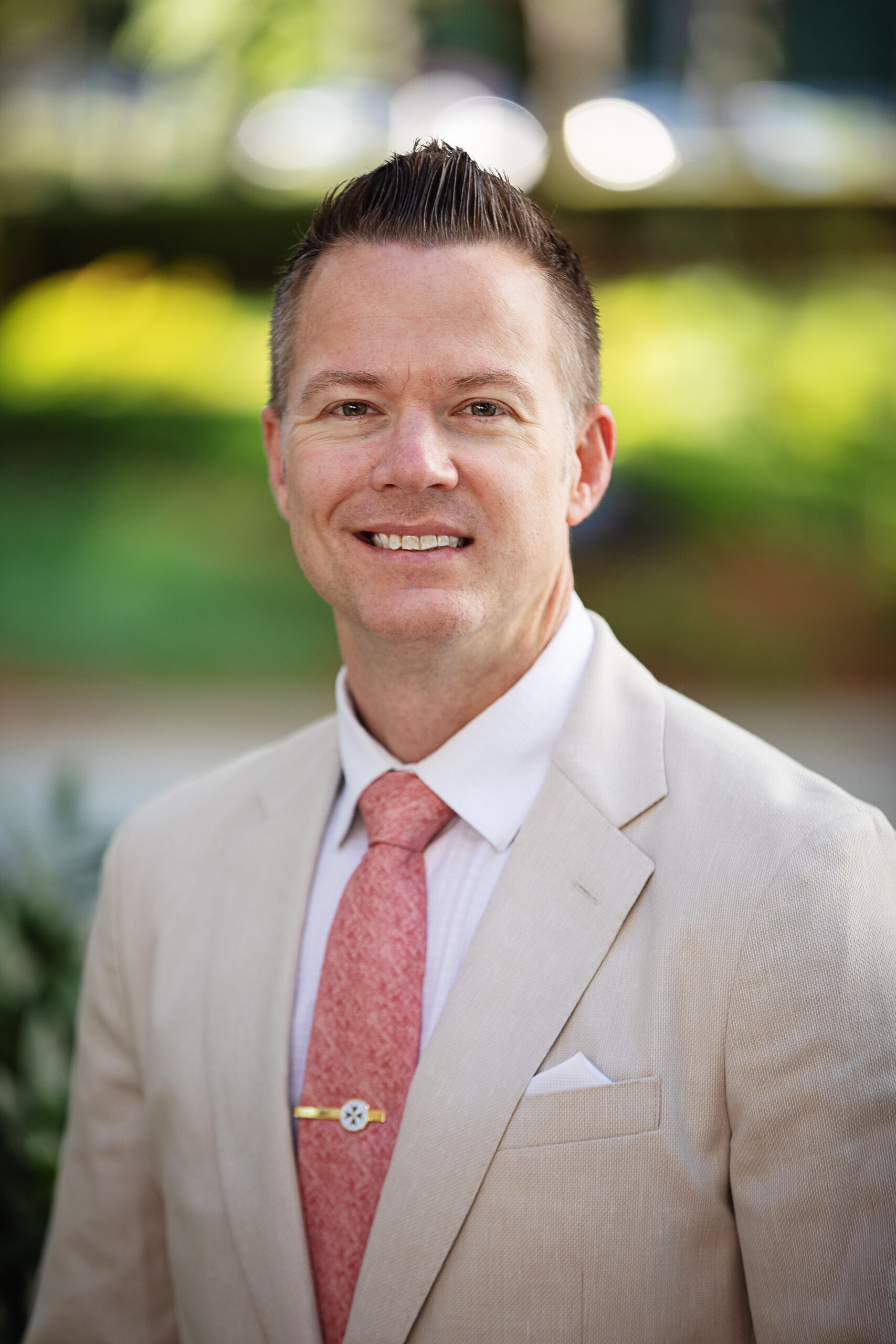 A man is smiling and wearing a light beige suit with a white shirt and a peach-colored tie. He has short, neatly styled hair. The background is outdoors with greenery and blurred elements.