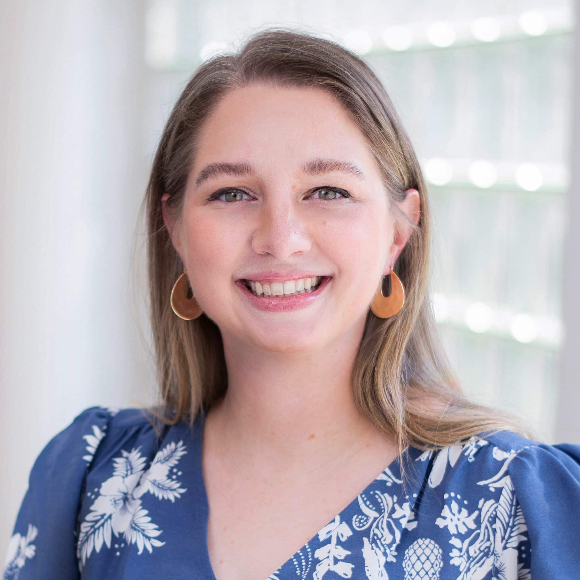 A woman with long, light brown hair smiles at the camera. She is wearing a blue dress with a white floral pattern and gold hoop earrings. The background features a window with diffused light.