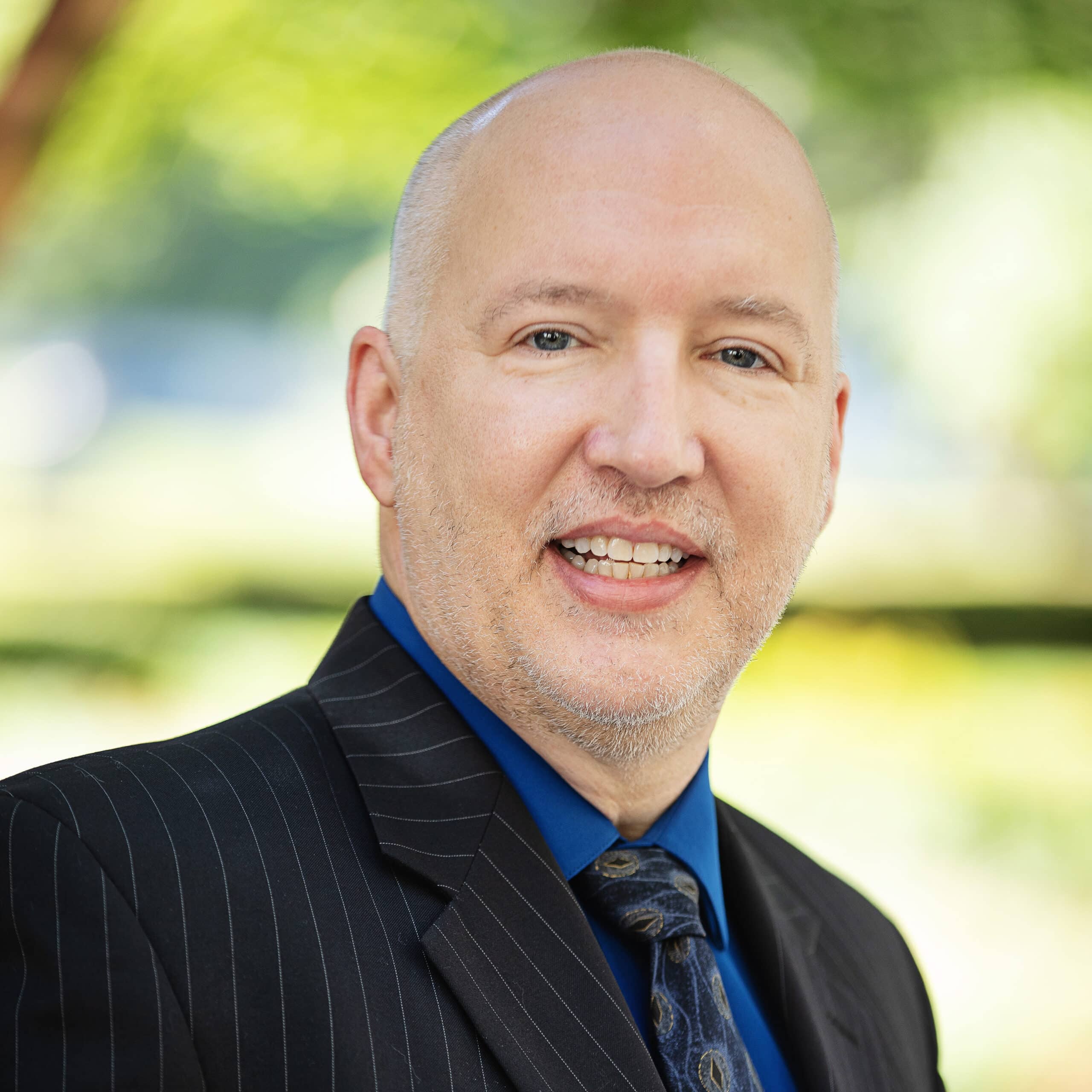 A man with a light beard is smiling at the camera. He is wearing a dark pinstripe suit, a blue dress shirt, and a patterned tie. The background is blurred greenery.