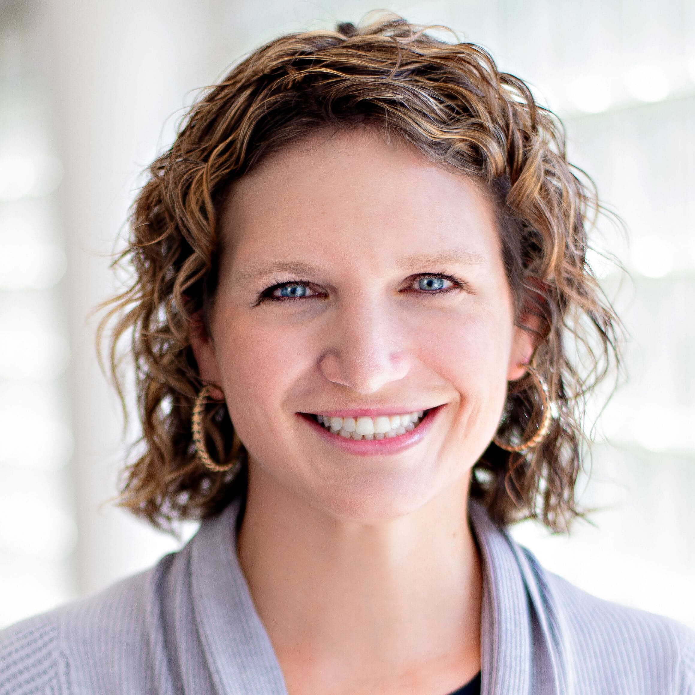 A smiling woman with wavy, shoulder-length brown hair is shown in a well-lit indoor setting. She is wearing a light gray sweater and hoop earrings. The background appears to be softly blurred, giving focus to her face.