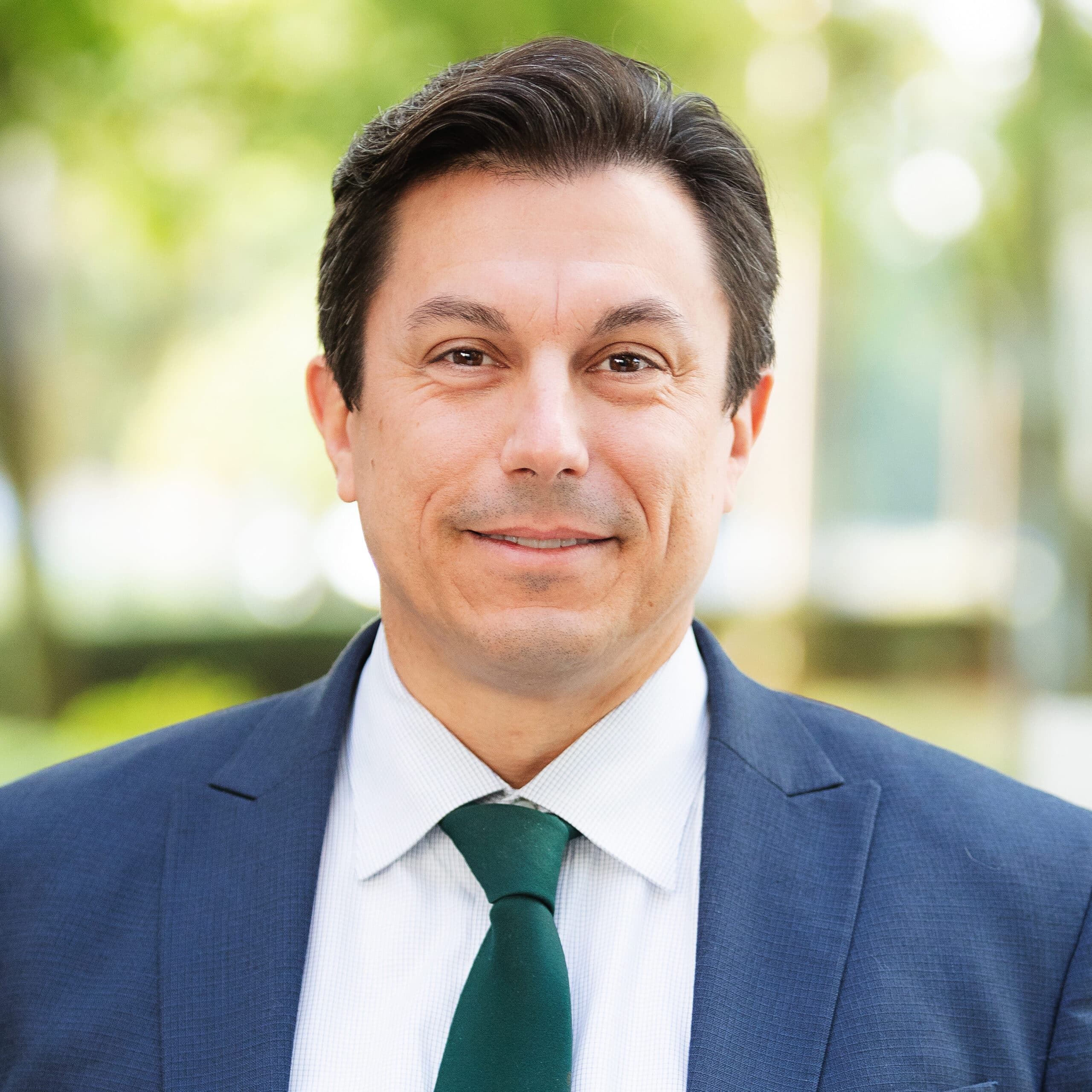 A person wearing a navy blue suit, white shirt, and green tie is smiling while standing outdoors.