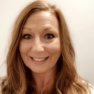 A woman with long, wavy, light brown hair is smiling at the camera. She is wearing a light-colored top and a necklace, with a plain light background behind her.