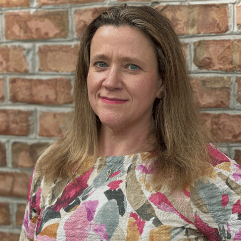 A woman with long brown hair smiles while standing in front of a brick wall. She is wearing a textured, long-sleeve blouse with a colorful floral pattern in shades of pink, orange, and green.