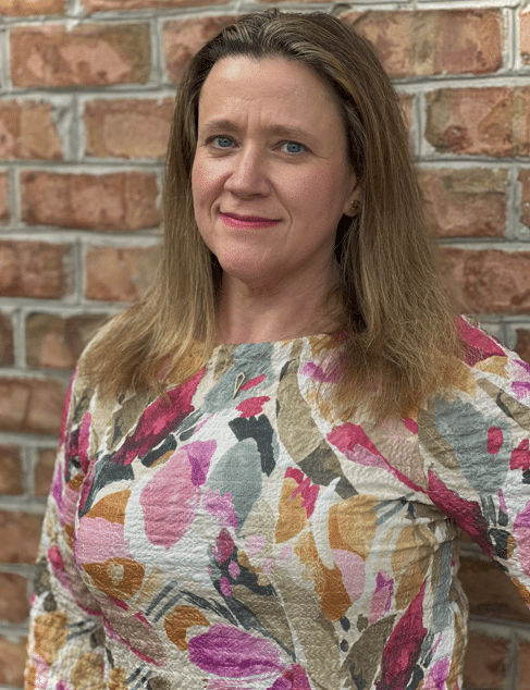 A woman with long brown hair smiles while standing in front of a brick wall. She is wearing a textured, long-sleeve blouse with a colorful floral pattern in shades of pink, orange, and green.
