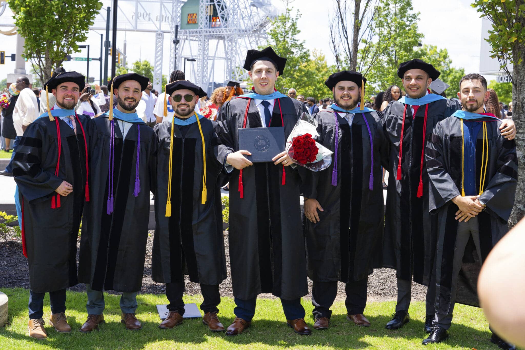 Seven graduates in caps and gowns smile and pose together outdoors on a sunny day. One holds a diploma and bouquet of roses. People and greenery can be seen in the background.