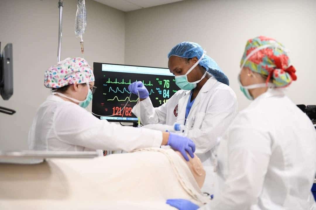 Three medical professionals in scrubs and masks work together in a hospital room, monitoring a patient’s vital signs displayed on a screen behind them.