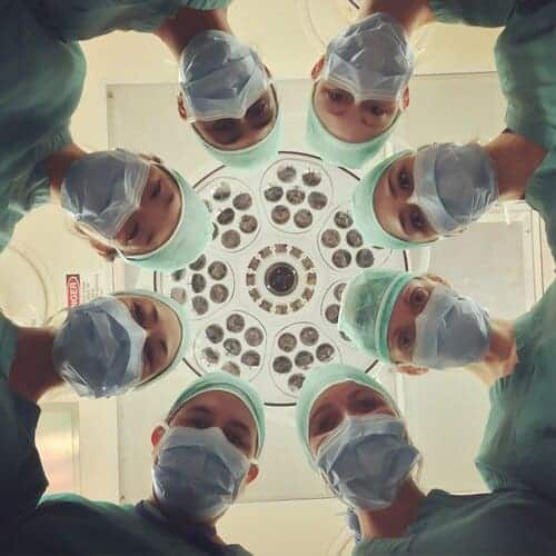 A group of surgeons wearing scrubs and masks stand in a circle under an operating room light, looking down toward the camera.
