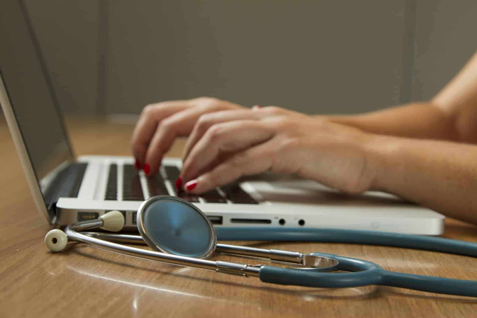 Person with red nail polish typing on a laptop, with a stethoscope resting on the wooden table in front of them.