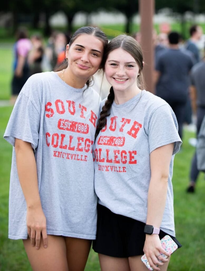 Two young women smile and pose together outdoors, both wearing matching gray “South College Knoxville” T-shirts. One holds a phone. People and greenery can be seen in the blurred background.